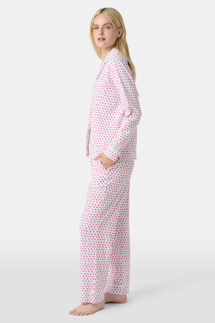 A woman stands barefoot in Roller Rabbit's Hearts Long Sleeve Polo Pajamas, a light pink Pima cotton set with a collared button-up top and matching pants, both decorated with small pink and purple hearts.