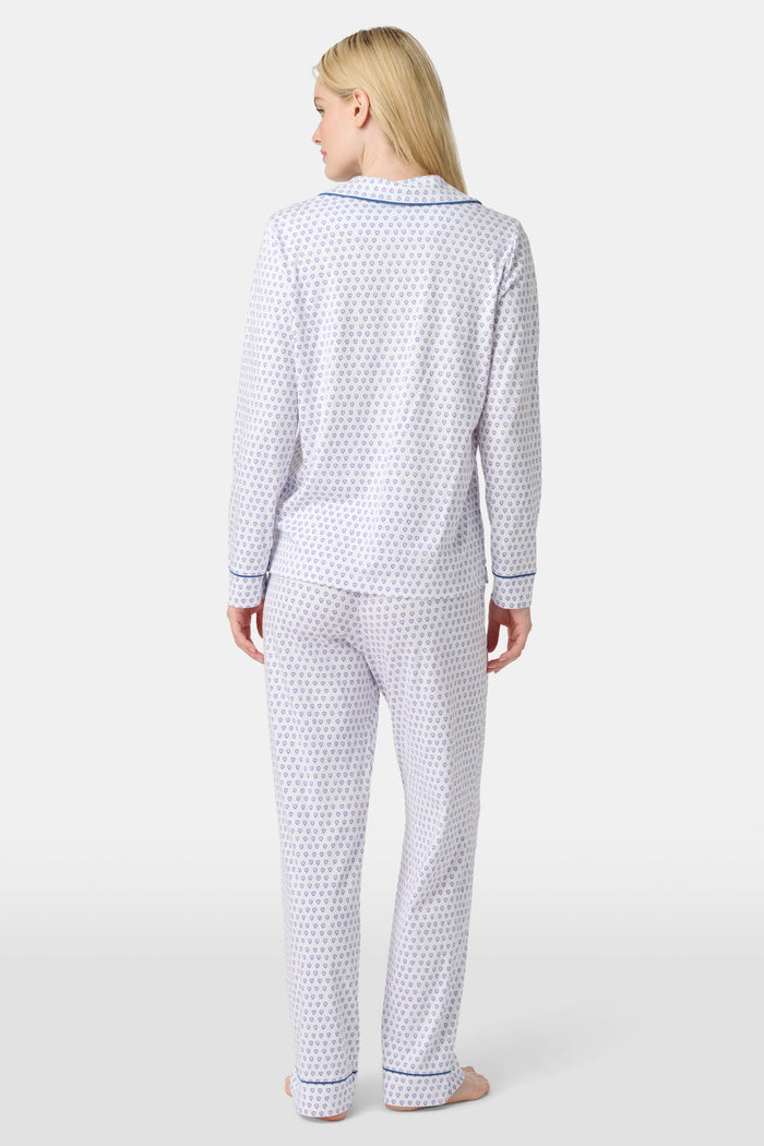 A woman stands barefoot in Roller Rabbit’s Hearts Long Sleeve Polo Pajamas, a Pima cotton set featuring a button-up top and long pants with blue piping and small blue heart patterns on a white background.