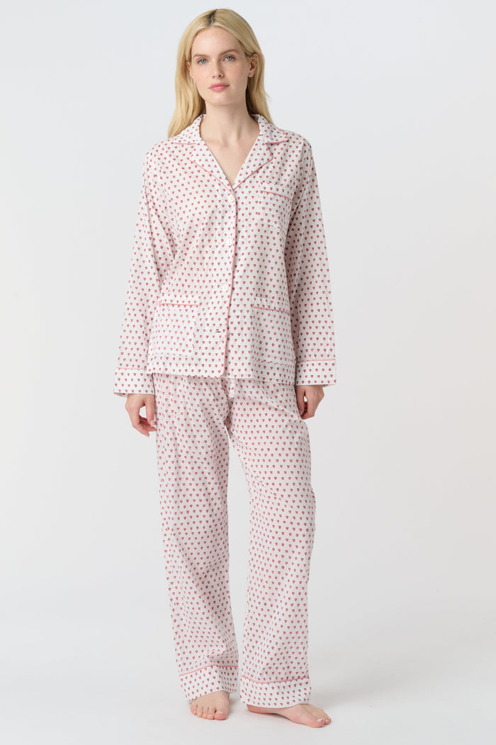 A woman stands barefoot in the Roller Rabbit Hearts Loungewear, a cotton pajama set with a button-up collared top and loose pants. The set features a pink and blue heart print. The background is plain white.