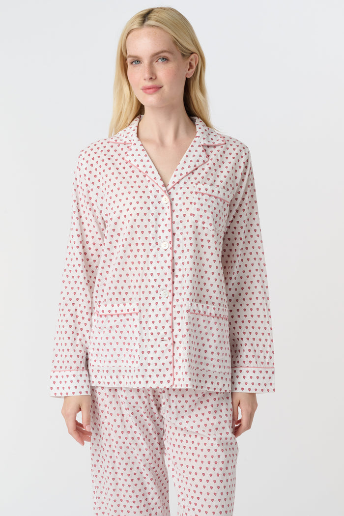 A woman stands barefoot in the Roller Rabbit Hearts Loungewear, a cotton pajama set with a button-up collared top and loose pants. The set features a pink and blue heart print. The background is plain white.