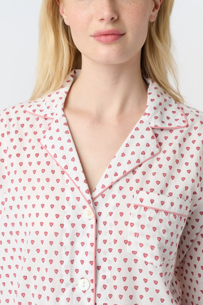 A woman stands barefoot in the Roller Rabbit Hearts Loungewear, a cotton pajama set with a button-up collared top and loose pants. The set features a pink and blue heart print. The background is plain white.