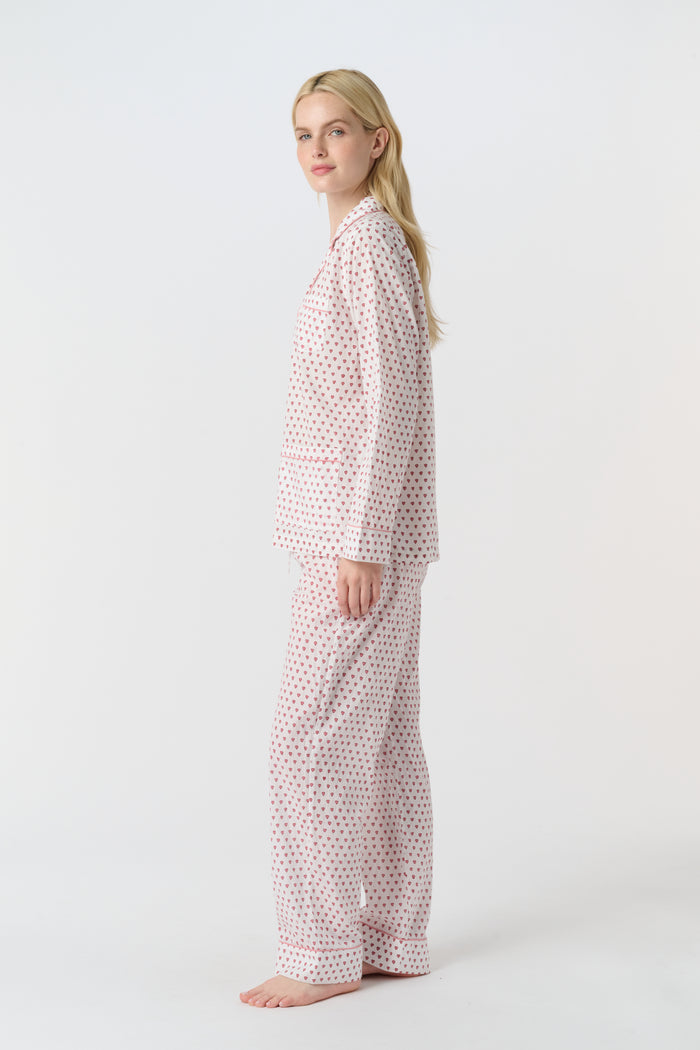 A woman stands barefoot in the Roller Rabbit Hearts Loungewear, a cotton pajama set with a button-up collared top and loose pants. The set features a pink and blue heart print. The background is plain white.