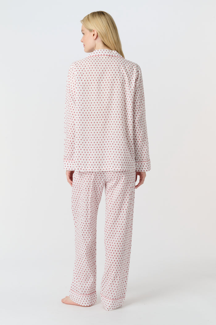 A woman stands barefoot in the Roller Rabbit Hearts Loungewear, a cotton pajama set with a button-up collared top and loose pants. The set features a pink and blue heart print. The background is plain white.