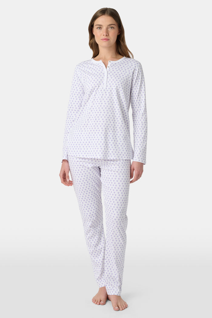 A woman stands barefoot in Roller Rabbit's Hearts Pajamas—a white, long-sleeve cotton set with a light lavender heart pattern, a round buttoned neckline top, and full-length pants—posed against a plain white background.