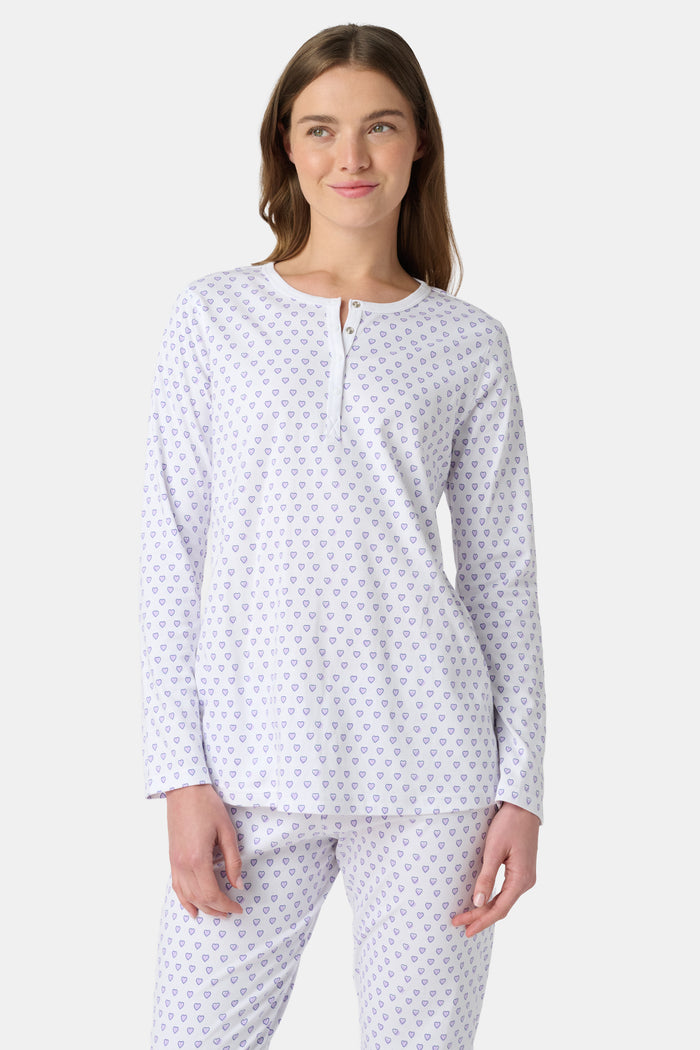 A woman stands barefoot in Roller Rabbit's Hearts Pajamas—a white, long-sleeve cotton set with a light lavender heart pattern, a round buttoned neckline top, and full-length pants—posed against a plain white background.