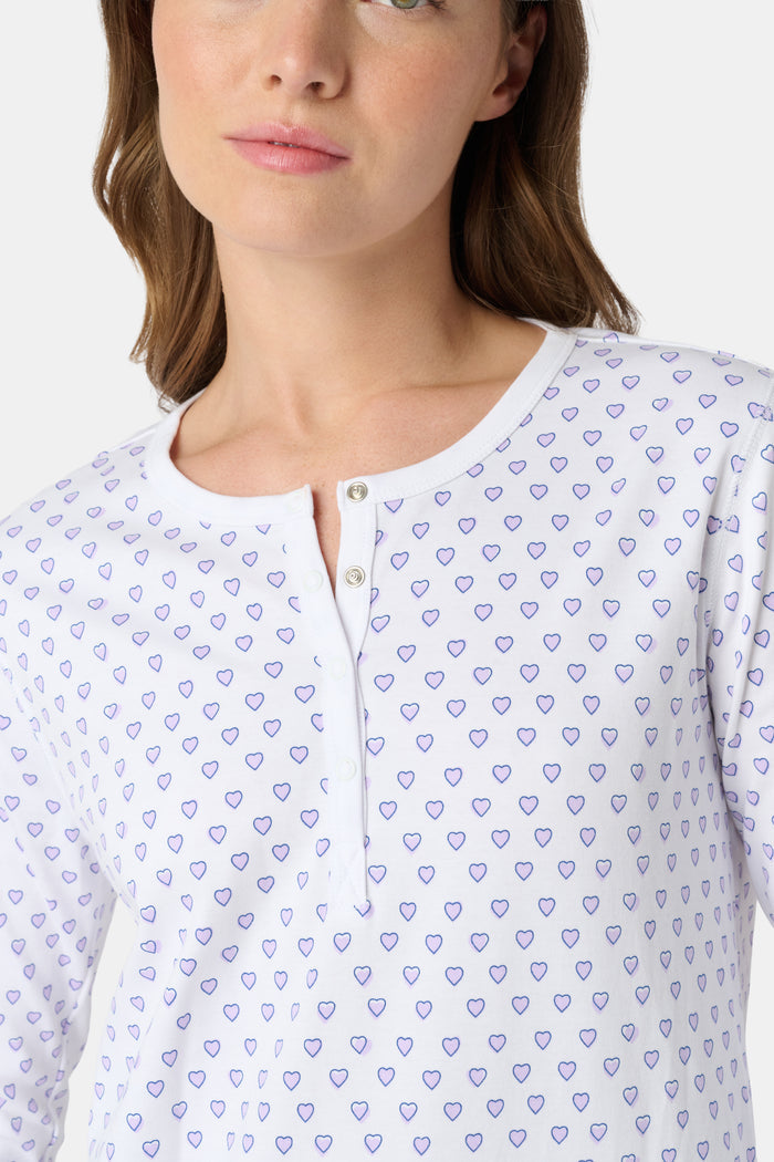 A woman stands barefoot in Roller Rabbit's Hearts Pajamas—a white, long-sleeve cotton set with a light lavender heart pattern, a round buttoned neckline top, and full-length pants—posed against a plain white background.
