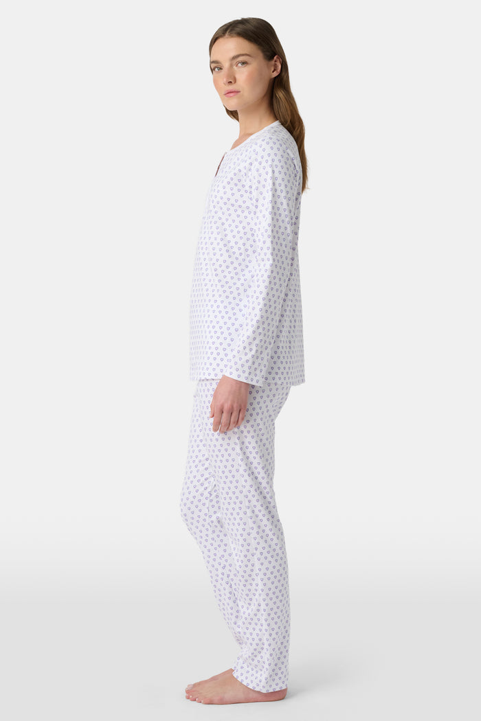A woman stands barefoot in Roller Rabbit's Hearts Pajamas—a white, long-sleeve cotton set with a light lavender heart pattern, a round buttoned neckline top, and full-length pants—posed against a plain white background.
