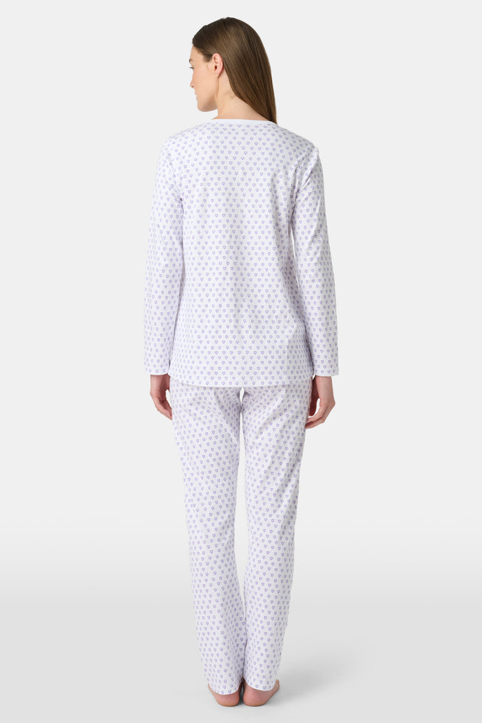 A woman stands barefoot in Roller Rabbit's Hearts Pajamas—a white, long-sleeve cotton set with a light lavender heart pattern, a round buttoned neckline top, and full-length pants—posed against a plain white background.