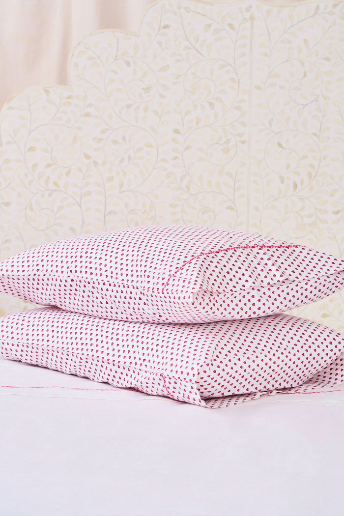 Two Hearts Pillowcase Set by Roller Rabbit, in white with small red dots and hearts print, are stacked on a white bed against a cream floral-patterned wall.