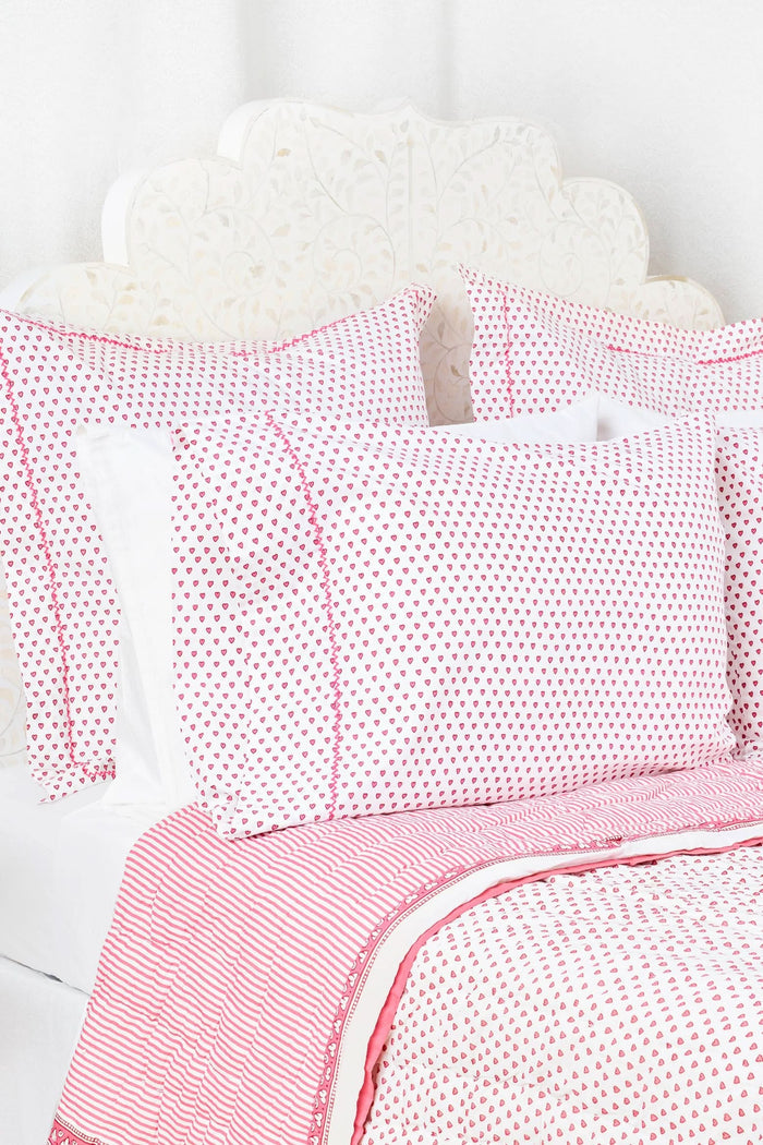 Two Hearts Pillowcase Set by Roller Rabbit, in white with small red dots and hearts print, are stacked on a white bed against a cream floral-patterned wall.
