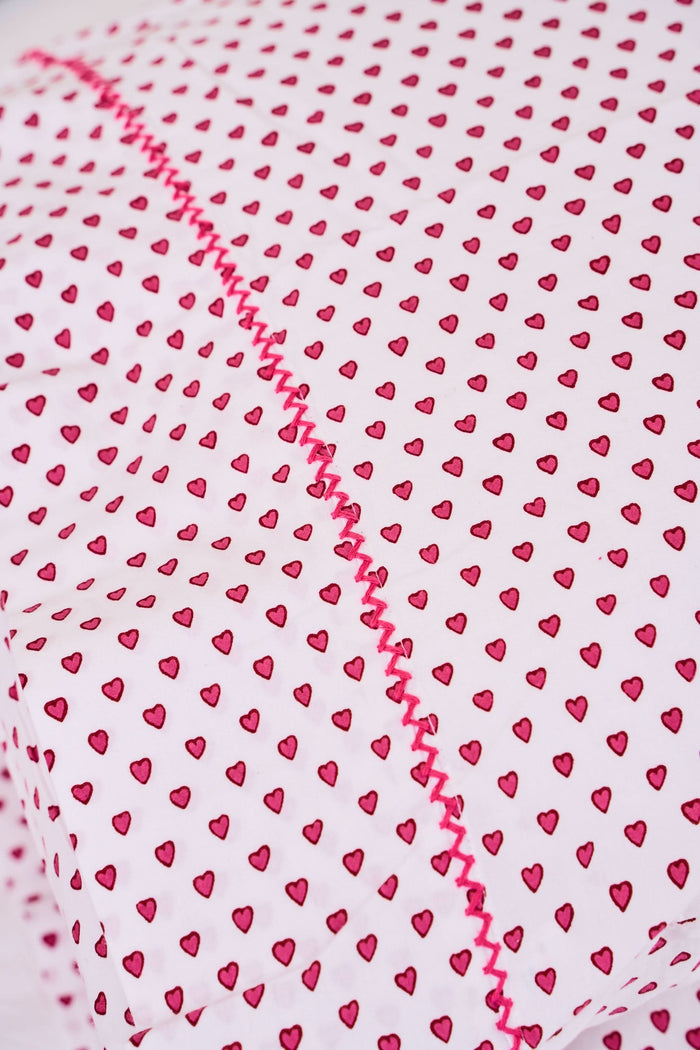 Two Hearts Pillowcase Set by Roller Rabbit, in white with small red dots and hearts print, are stacked on a white bed against a cream floral-patterned wall.