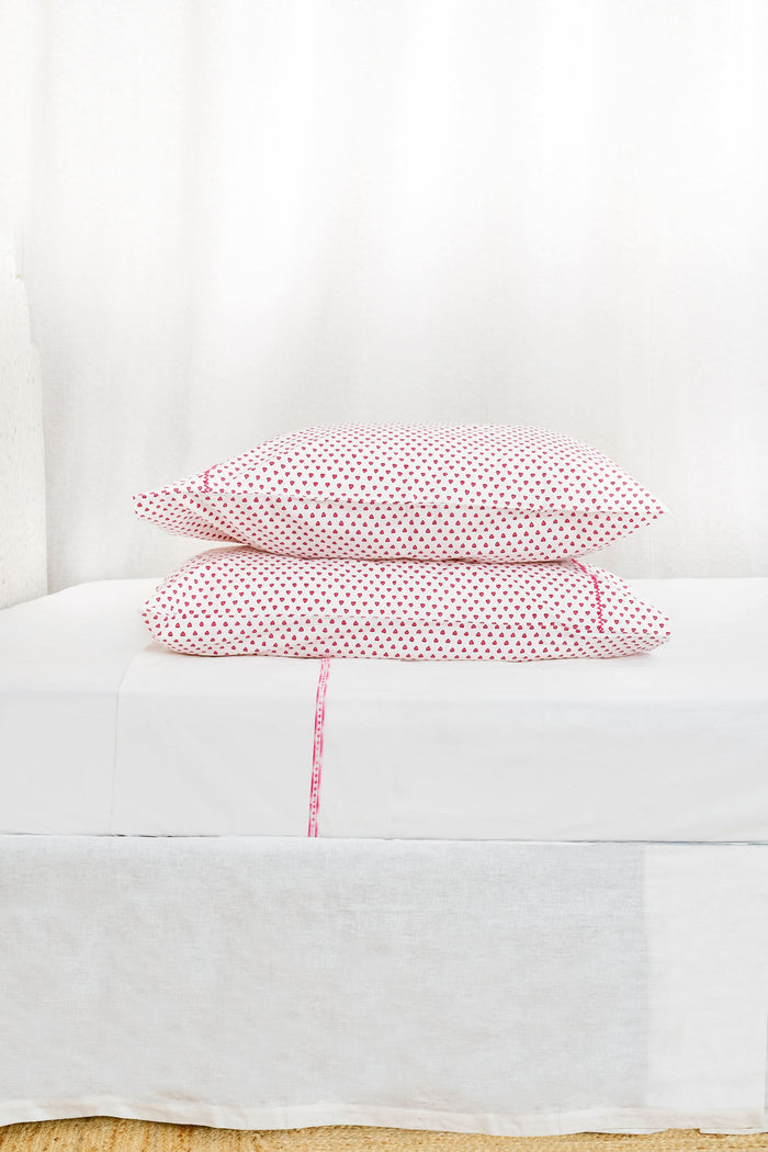 Two Hearts Pillowcase Set by Roller Rabbit, in white with small red dots and hearts print, are stacked on a white bed against a cream floral-patterned wall.