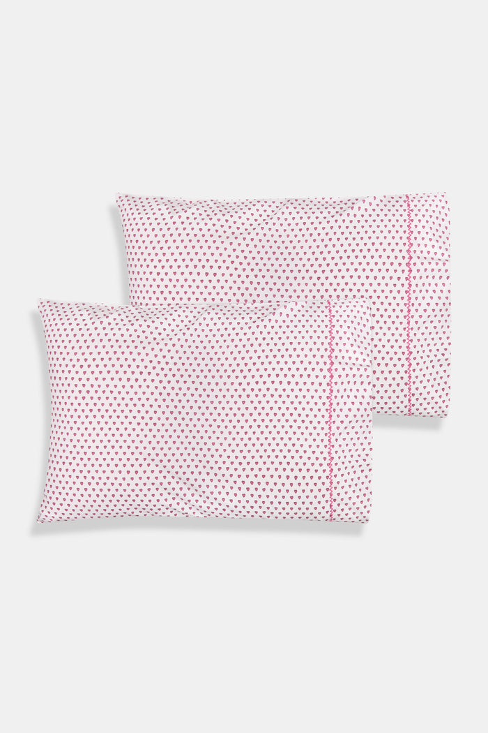 Two Hearts Pillowcase Set by Roller Rabbit, in white with small red dots and hearts print, are stacked on a white bed against a cream floral-patterned wall.