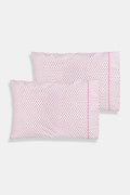 Two Hearts Pillowcase Set by Roller Rabbit, in white with small red dots and hearts print, are stacked on a white bed against a cream floral-patterned wall.