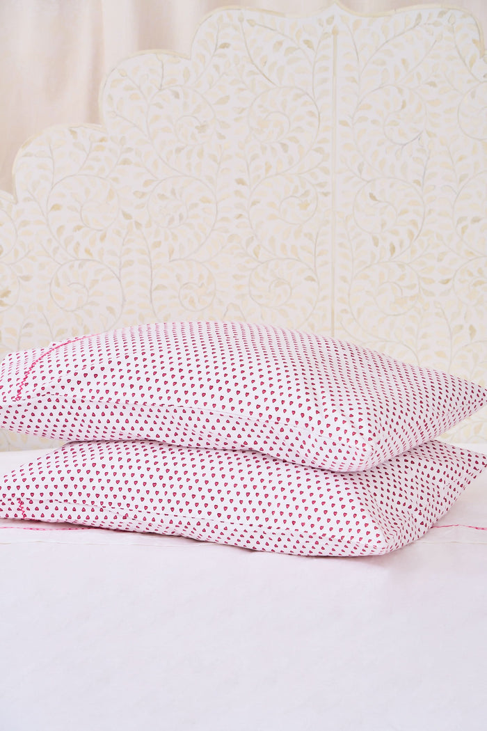 Two Hearts Pillowcase Set by Roller Rabbit, in white with small red dots and hearts print, are stacked on a white bed against a cream floral-patterned wall.