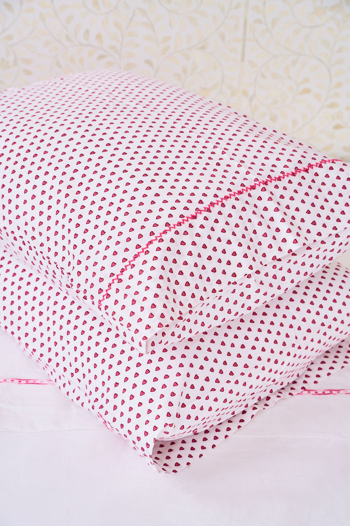 Two Hearts Pillowcase Set by Roller Rabbit, in white with small red dots and hearts print, are stacked on a white bed against a cream floral-patterned wall.