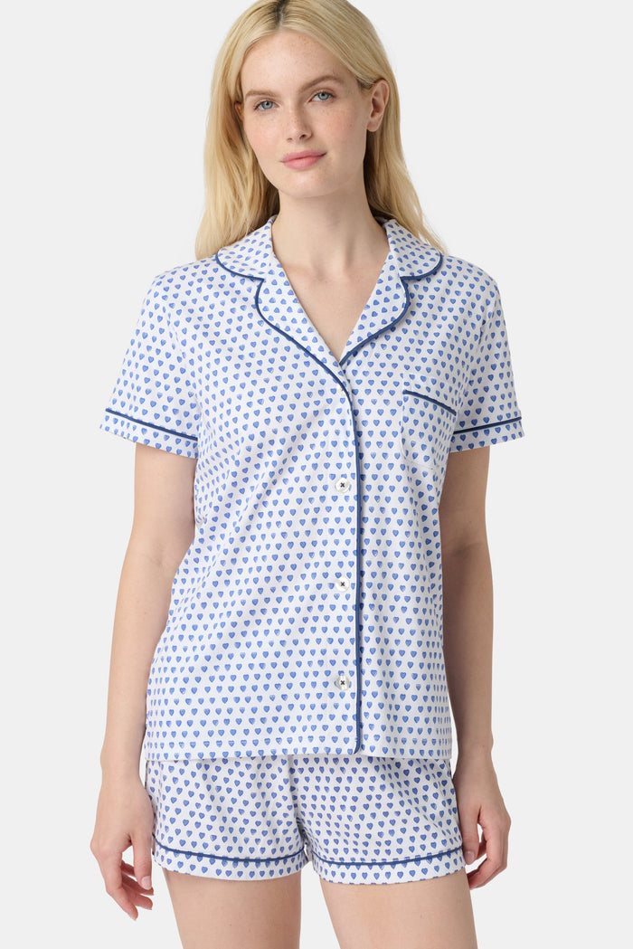 A woman with long blonde hair wears Roller Rabbit Hearts Polo Pajamas, a 100 Pima cotton set made in Peru with a blue heart print, short sleeves, blue trim, and shorts. She stands smiling gently against a plain backdrop.