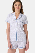 A woman wears the Roller Rabbit Hearts Polo Pajamas, made from 100 cotton with a white base and small heart print. The set features a short-sleeve, button-up collared top with chest pocket and matching shorts. She stands facing forward against a plain background.