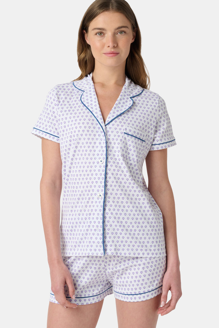 A woman wears the Roller Rabbit Hearts Polo Pajamas, made from 100 cotton with a white base and small heart print. The set features a short-sleeve, button-up collared top with chest pocket and matching shorts. She stands facing forward against a plain background.