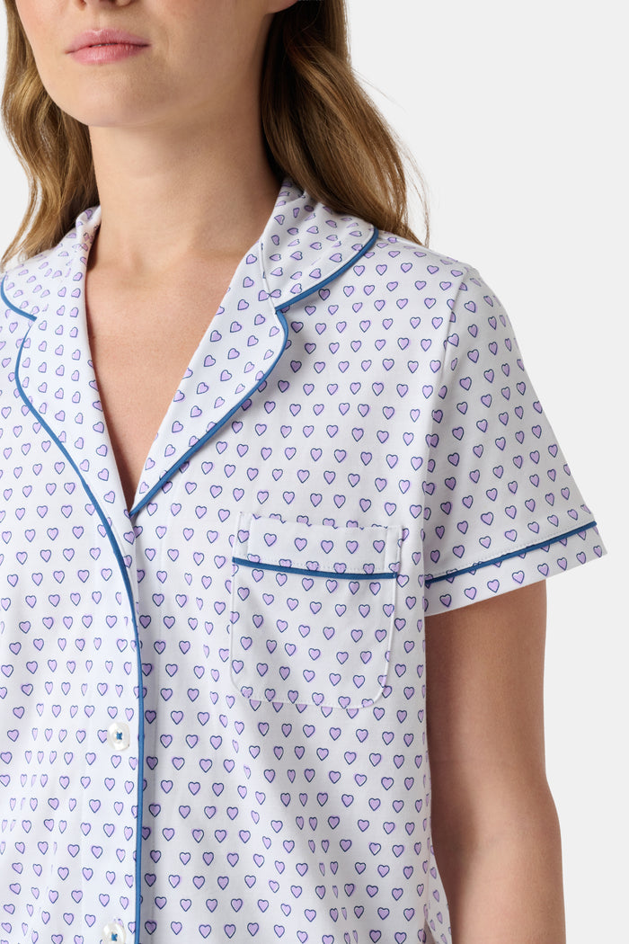A woman wears the Roller Rabbit Hearts Polo Pajamas, made from 100 cotton with a white base and small heart print. The set features a short-sleeve, button-up collared top with chest pocket and matching shorts. She stands facing forward against a plain background.