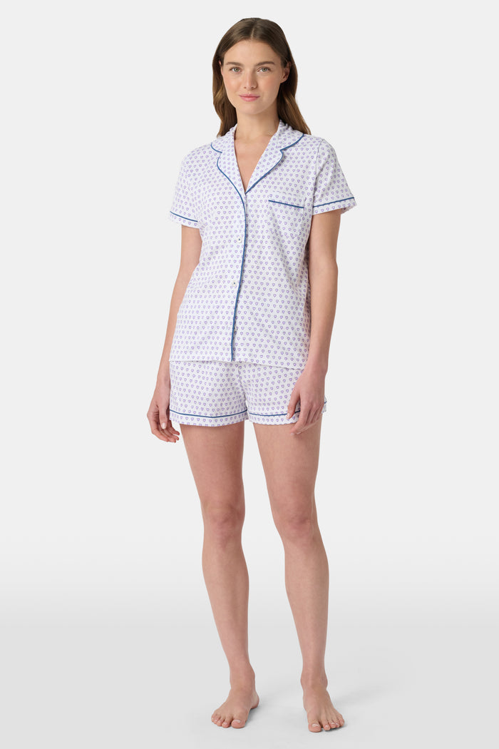 A woman wears the Roller Rabbit Hearts Polo Pajamas, made from 100 cotton with a white base and small heart print. The set features a short-sleeve, button-up collared top with chest pocket and matching shorts. She stands facing forward against a plain background.
