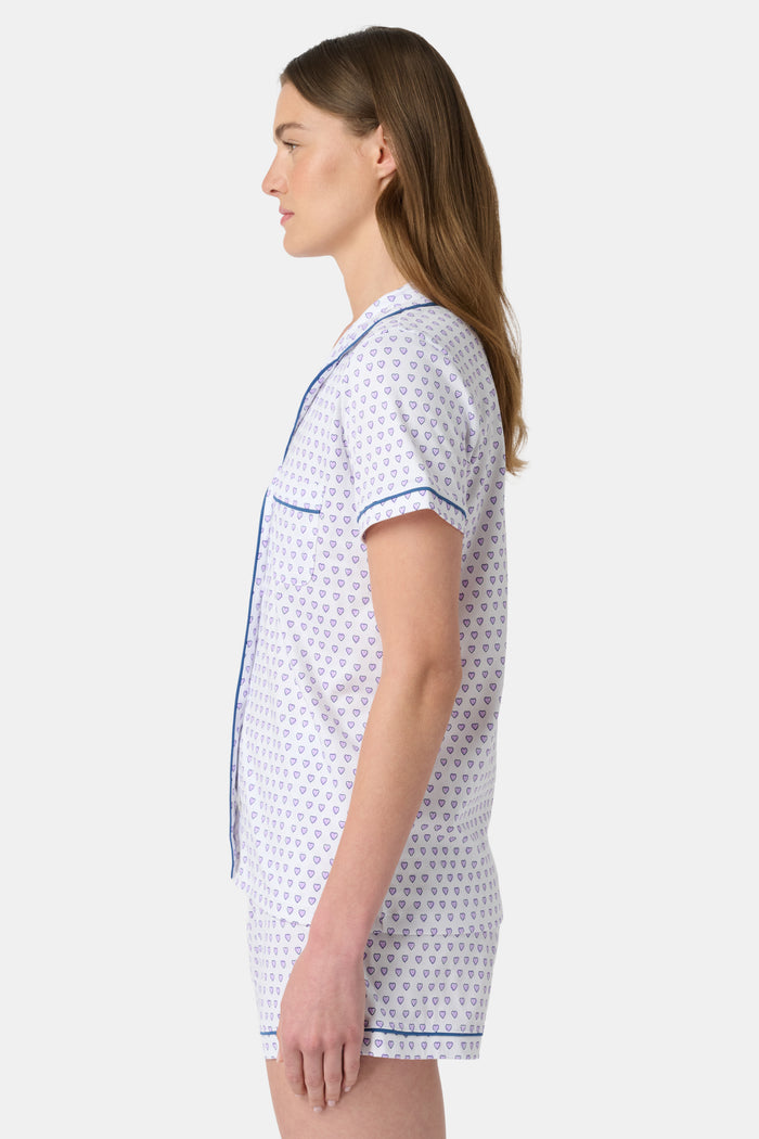 A woman wears the Roller Rabbit Hearts Polo Pajamas, made from 100 cotton with a white base and small heart print. The set features a short-sleeve, button-up collared top with chest pocket and matching shorts. She stands facing forward against a plain background.
