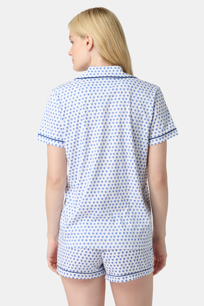 A woman with long blonde hair wears Roller Rabbit Hearts Polo Pajamas, a 100 Pima cotton set made in Peru with a blue heart print, short sleeves, blue trim, and shorts. She stands smiling gently against a plain backdrop.