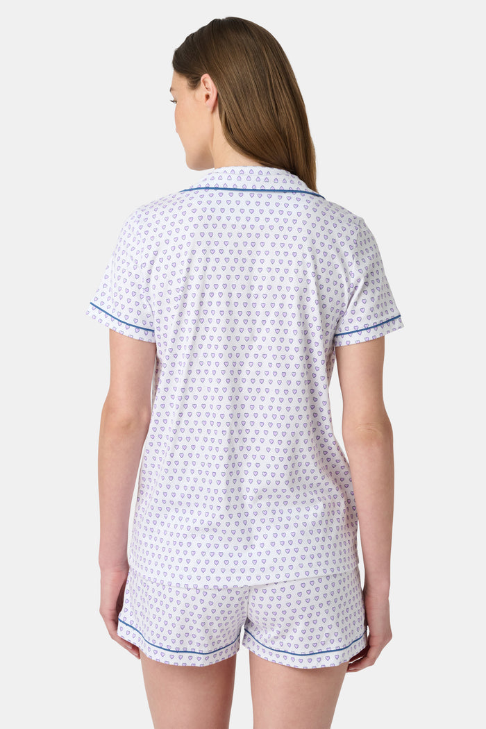 A woman wears the Roller Rabbit Hearts Polo Pajamas, made from 100 cotton with a white base and small heart print. The set features a short-sleeve, button-up collared top with chest pocket and matching shorts. She stands facing forward against a plain background.