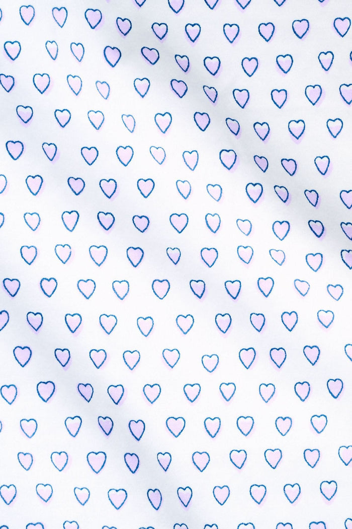 A woman wears the Roller Rabbit Hearts Polo Pajamas, made from 100 cotton with a white base and small heart print. The set features a short-sleeve, button-up collared top with chest pocket and matching shorts. She stands facing forward against a plain background.