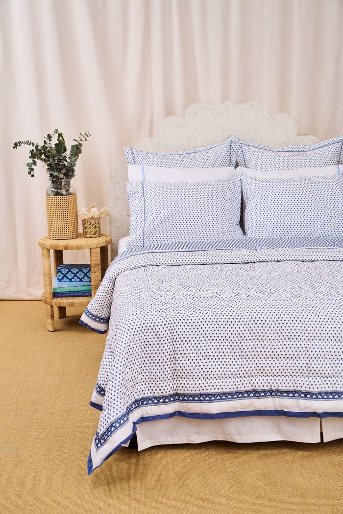 A neatly made bed features the Roller Rabbit Hearts Quilt, blue and white patterned bedding, four pillows, and a cotton voile quilt. A woven nightstand with a plant and blue towel stands beside it against a beige backdrop.