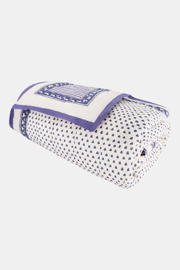 A neatly made bed features the Roller Rabbit Hearts Quilt, blue and white patterned bedding, four pillows, and a cotton voile quilt. A woven nightstand with a plant and blue towel stands beside it against a beige backdrop.