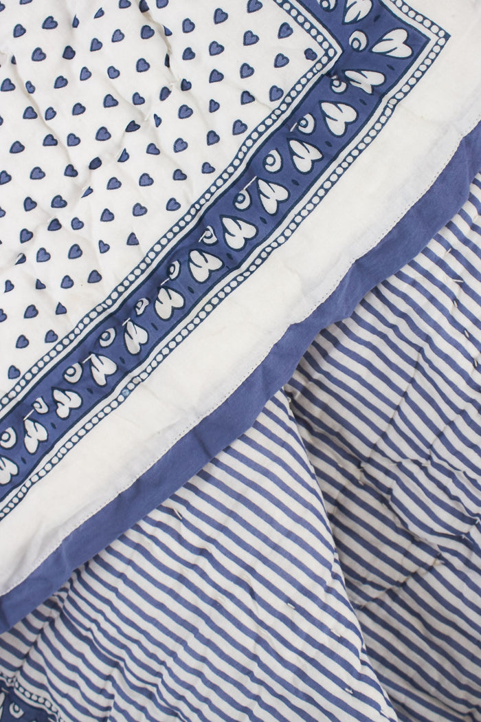 A neatly made bed features the Roller Rabbit Hearts Quilt, blue and white patterned bedding, four pillows, and a cotton voile quilt. A woven nightstand with a plant and blue towel stands beside it against a beige backdrop.