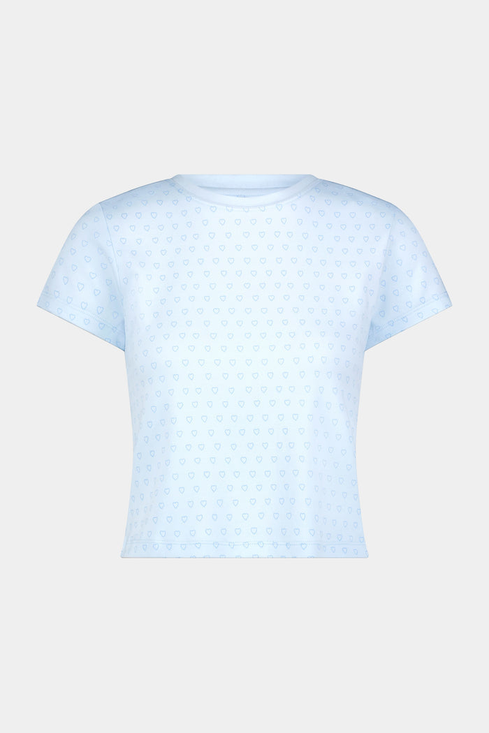 A woman with wavy dark hair gives a slight smile while wearing the Roller Rabbit Hearts Tonal Classic Baby Tee, standing against a plain white background.