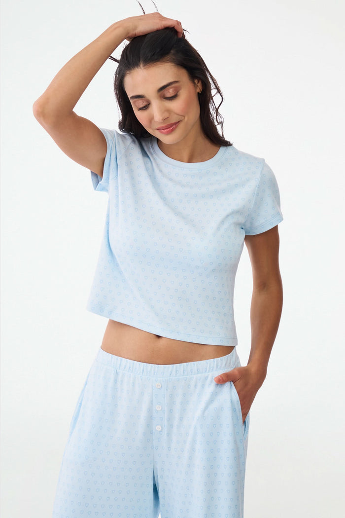 A woman with wavy dark hair gives a slight smile while wearing the Roller Rabbit Hearts Tonal Classic Baby Tee, standing against a plain white background.