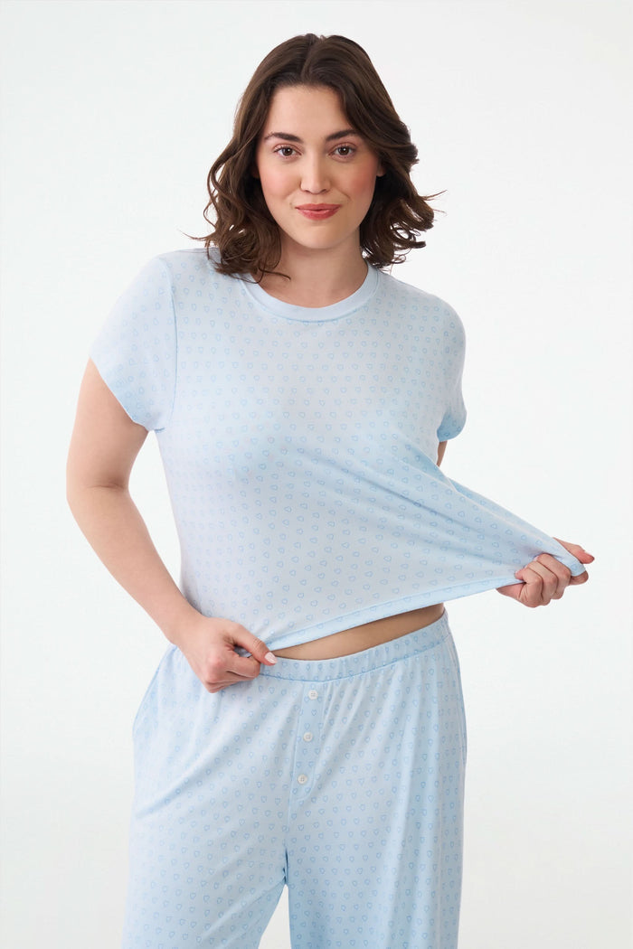 A woman with wavy dark hair gives a slight smile while wearing the Roller Rabbit Hearts Tonal Classic Baby Tee, standing against a plain white background.