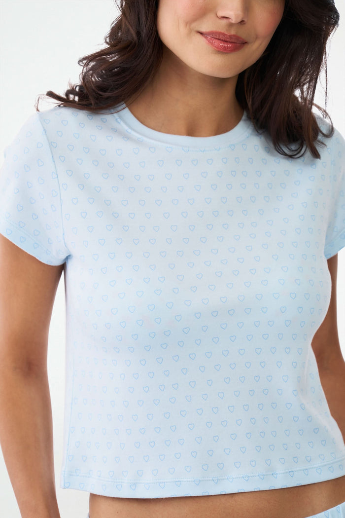 A woman with wavy dark hair gives a slight smile while wearing the Roller Rabbit Hearts Tonal Classic Baby Tee, standing against a plain white background.
