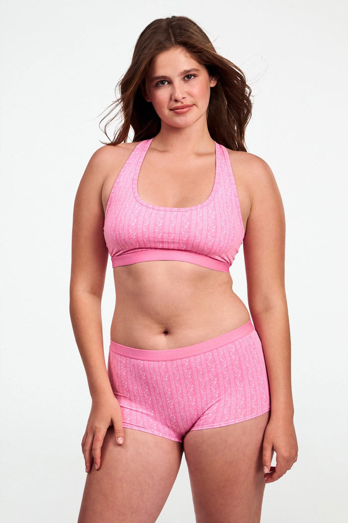 A woman poses against a plain background, wearing Roller Rabbit’s Hearts Vine Crosby Boyshort and a matching sports bra. She smiles softly at the camera, with one hand by her head.