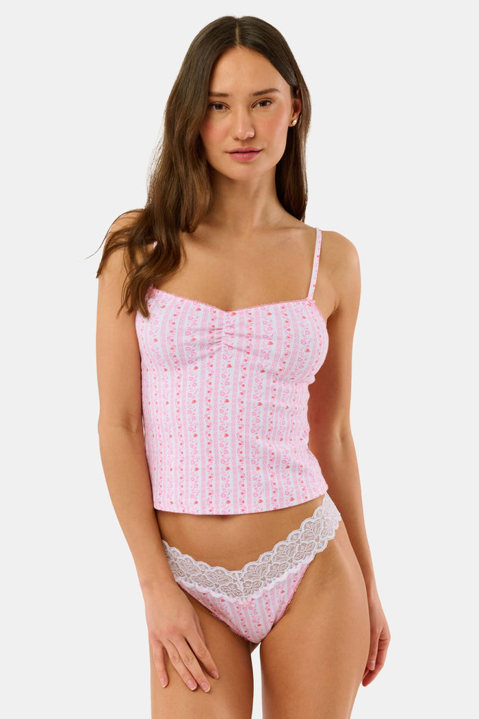 A woman wears the Hearts Vine Greene Cami pajama set by Roller Rabbit in pink and white with a small floral pattern, standing against a plain light background.
