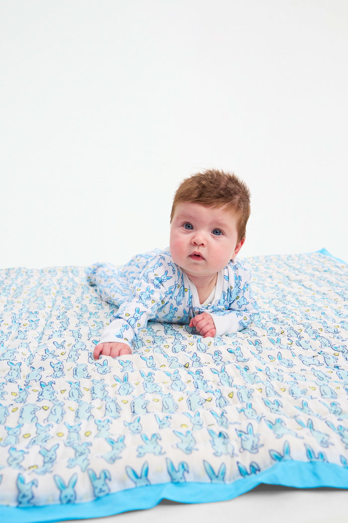 The Roller Rabbit Hoppy Baby Quilt features a playful blue and yellow Hoppy the Bunny pattern on white cotton, accented with a blue border and shown rolled up on a light surface.