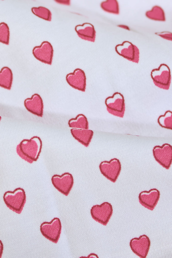 A baby with light skin and light brown hair lies on a pink and white striped blanket wearing the Roller Rabbit Infant Hearts Polo Onesie, looking slightly to the side.