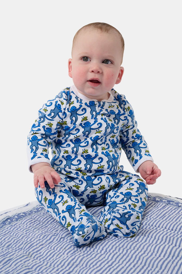 A baby sits on a blue and white striped blanket, wearing the Roller Rabbit Infant Monkey Zipper Footie—a premium cotton one-piece featuring a playful monkey print—while looking slightly upward with a happy expression.