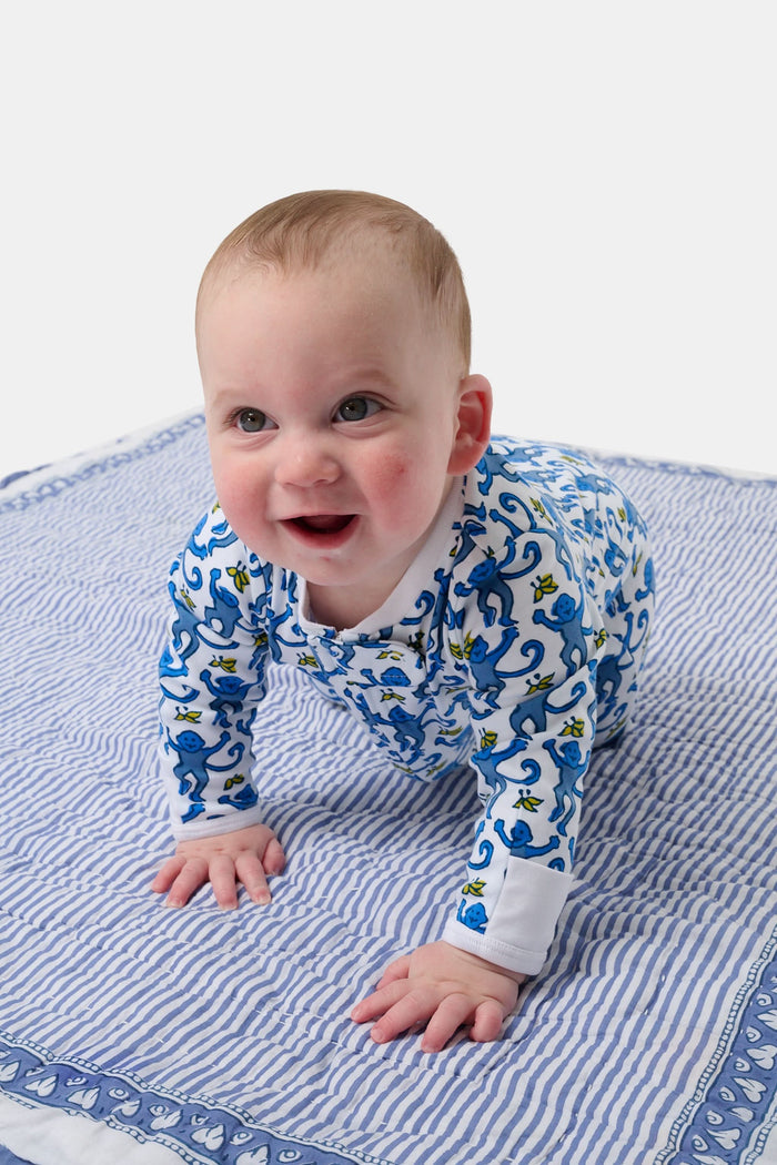 A baby sits on a blue and white striped blanket, wearing the Roller Rabbit Infant Monkey Zipper Footie—a premium cotton one-piece featuring a playful monkey print—while looking slightly upward with a happy expression.