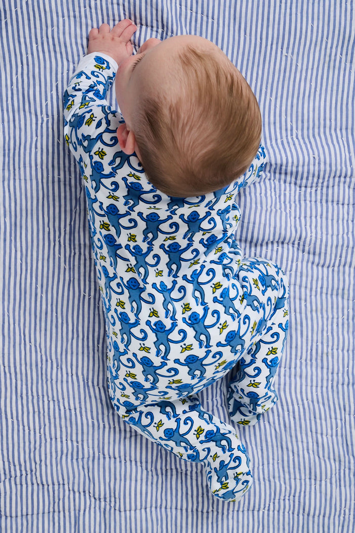 A baby sits on a blue and white striped blanket, wearing the Roller Rabbit Infant Monkey Zipper Footie—a premium cotton one-piece featuring a playful monkey print—while looking slightly upward with a happy expression.