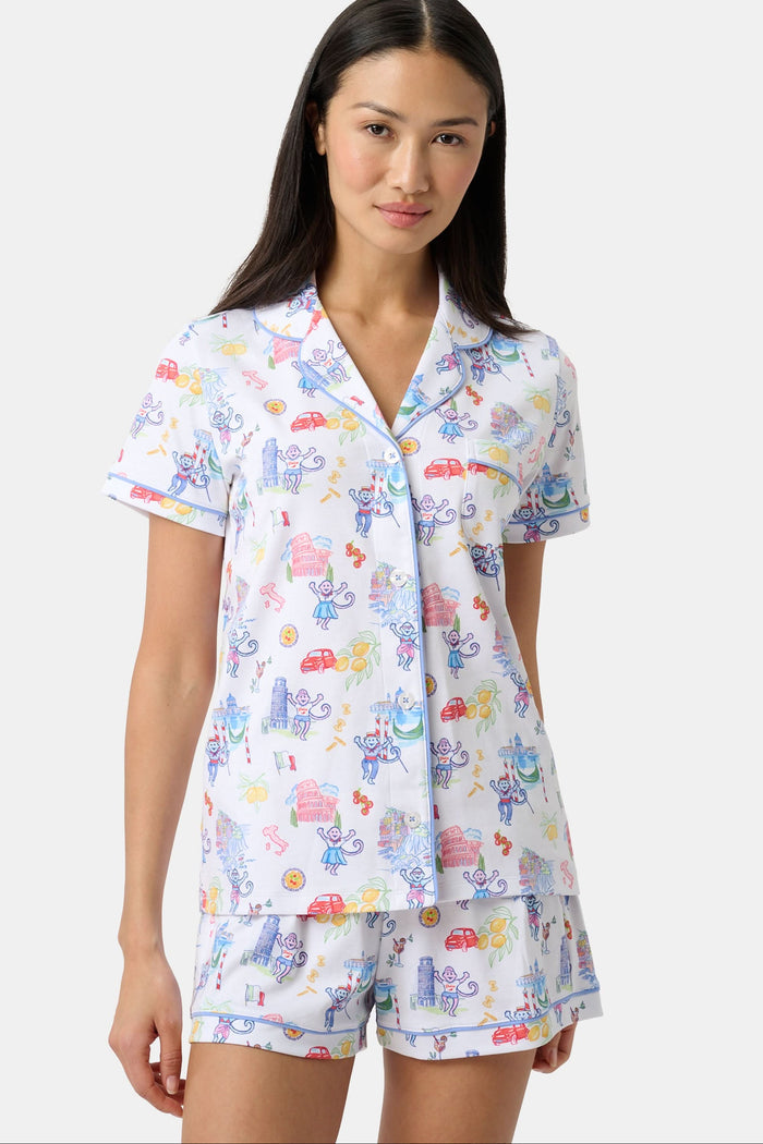 A woman with long dark hair wears Roller Rabbit’s Italy Monkey Polo Pajamas, a playful Pima Cotton set featuring cartoon animals and Italian motifs. The short-sleeved, button-up top pairs perfectly with the matching shorts.