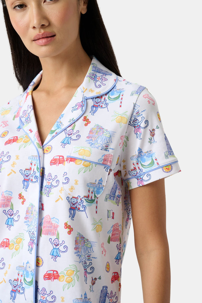 A woman with long dark hair wears Roller Rabbit’s Italy Monkey Polo Pajamas, a playful Pima Cotton set featuring cartoon animals and Italian motifs. The short-sleeved, button-up top pairs perfectly with the matching shorts.