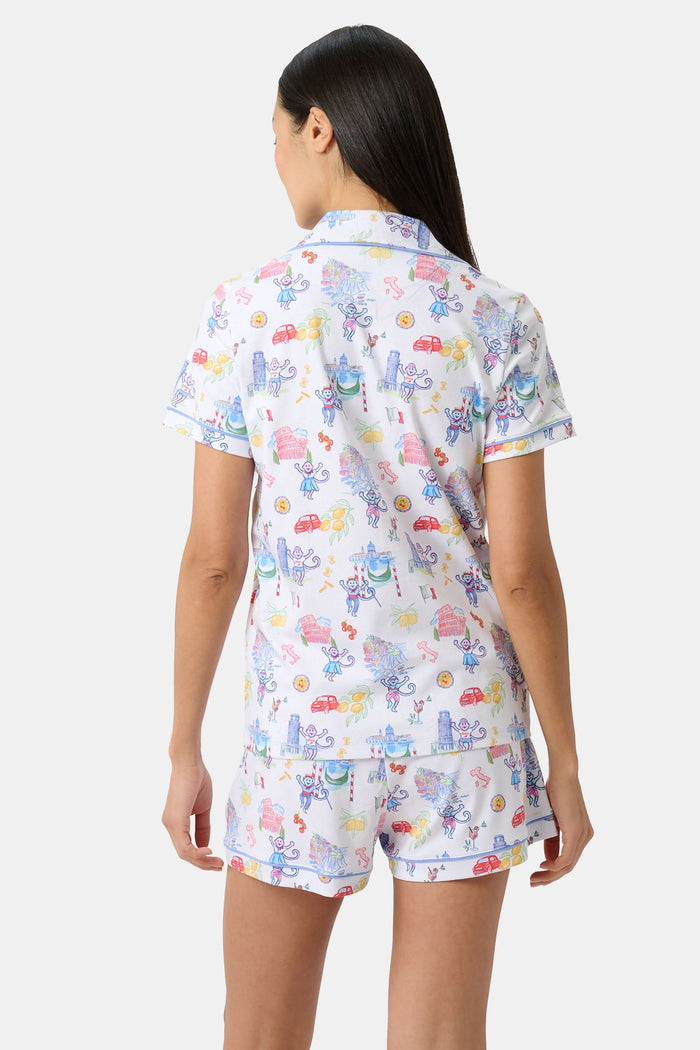 A woman with long dark hair wears Roller Rabbit’s Italy Monkey Polo Pajamas, a playful Pima Cotton set featuring cartoon animals and Italian motifs. The short-sleeved, button-up top pairs perfectly with the matching shorts.