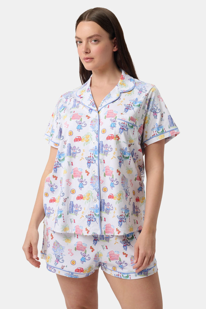 A woman with long dark hair wears Roller Rabbit’s Italy Monkey Polo Pajamas, a playful Pima Cotton set featuring cartoon animals and Italian motifs. The short-sleeved, button-up top pairs perfectly with the matching shorts.