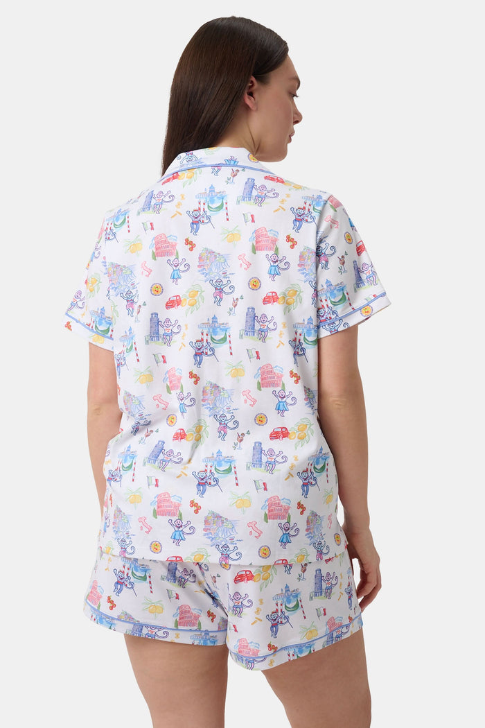 A woman with long dark hair wears Roller Rabbit’s Italy Monkey Polo Pajamas, a playful Pima Cotton set featuring cartoon animals and Italian motifs. The short-sleeved, button-up top pairs perfectly with the matching shorts.