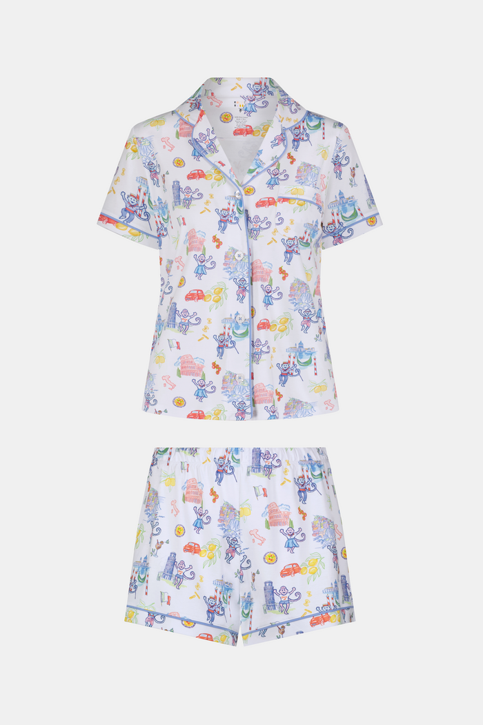 A woman with long dark hair wears Roller Rabbit’s Italy Monkey Polo Pajamas, a playful Pima Cotton set featuring cartoon animals and Italian motifs. The short-sleeved, button-up top pairs perfectly with the matching shorts.