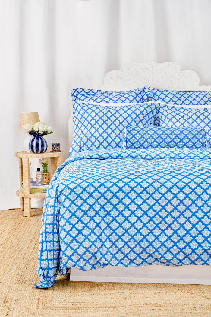 A neatly made bed features the Roller Rabbit Jemina Duvet Cover in blue and white, set before a white curtain. A small bedside table holds a striped vase with flowers, books, and a lamp atop a woven rug.